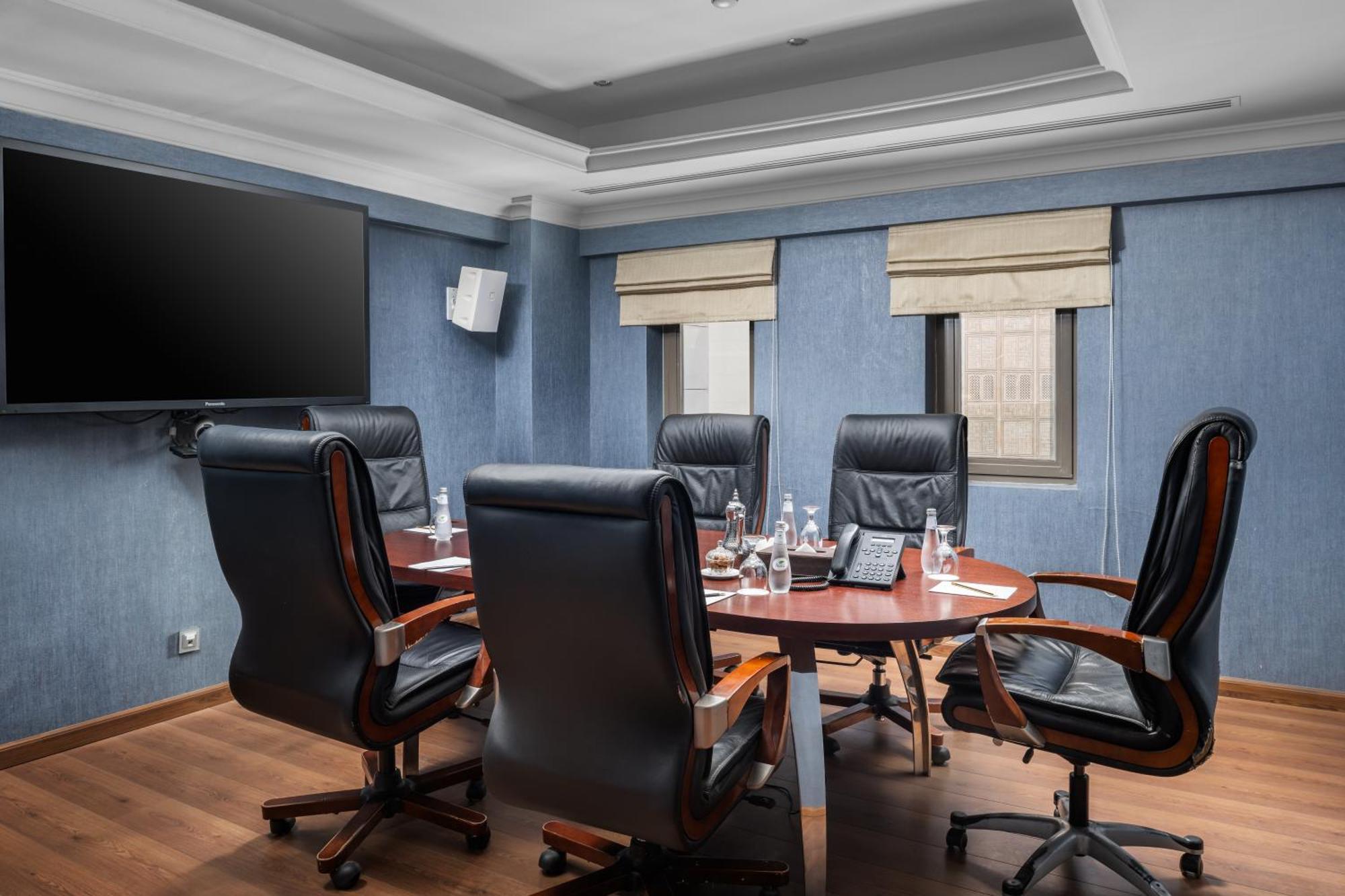 Al Aqeeq Madinah Hotel Медина Экстерьер фото The photo shows a modern conference room. It features a round wooden table surrounded by several black leather swivel chairs. The walls have a blue color scheme, and there is a large flat-screen TV mounted on one wall. The room has two windows with b