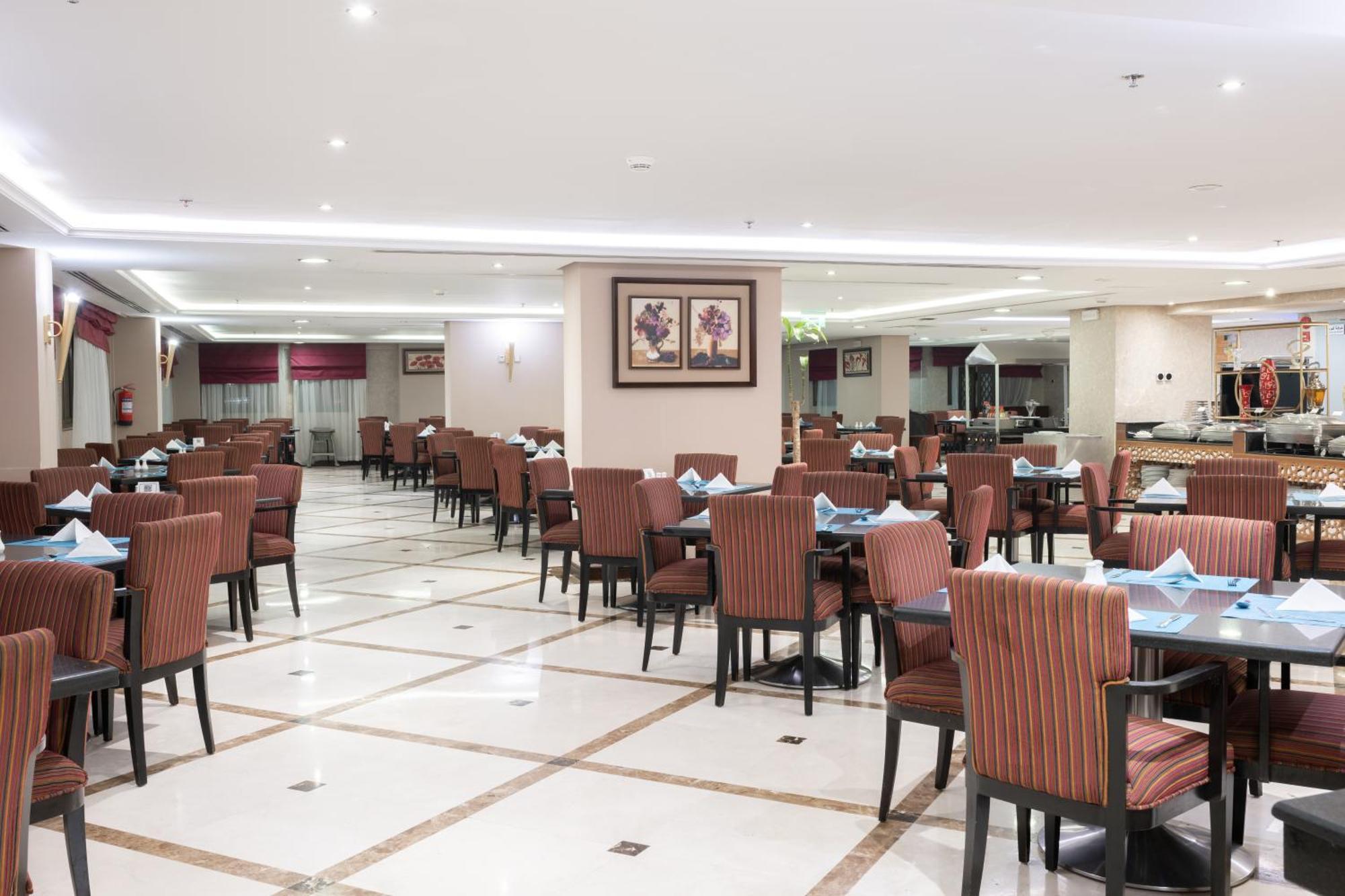 Al Aqeeq Madinah Hotel Медина Экстерьер фото The photo shows the interior of a spacious restaurant. It features a well-organized dining area with neatly arranged tables and chairs. The tables are dressed with blue napkins, and there is a light-colored flooring with a grid pattern. The walls are