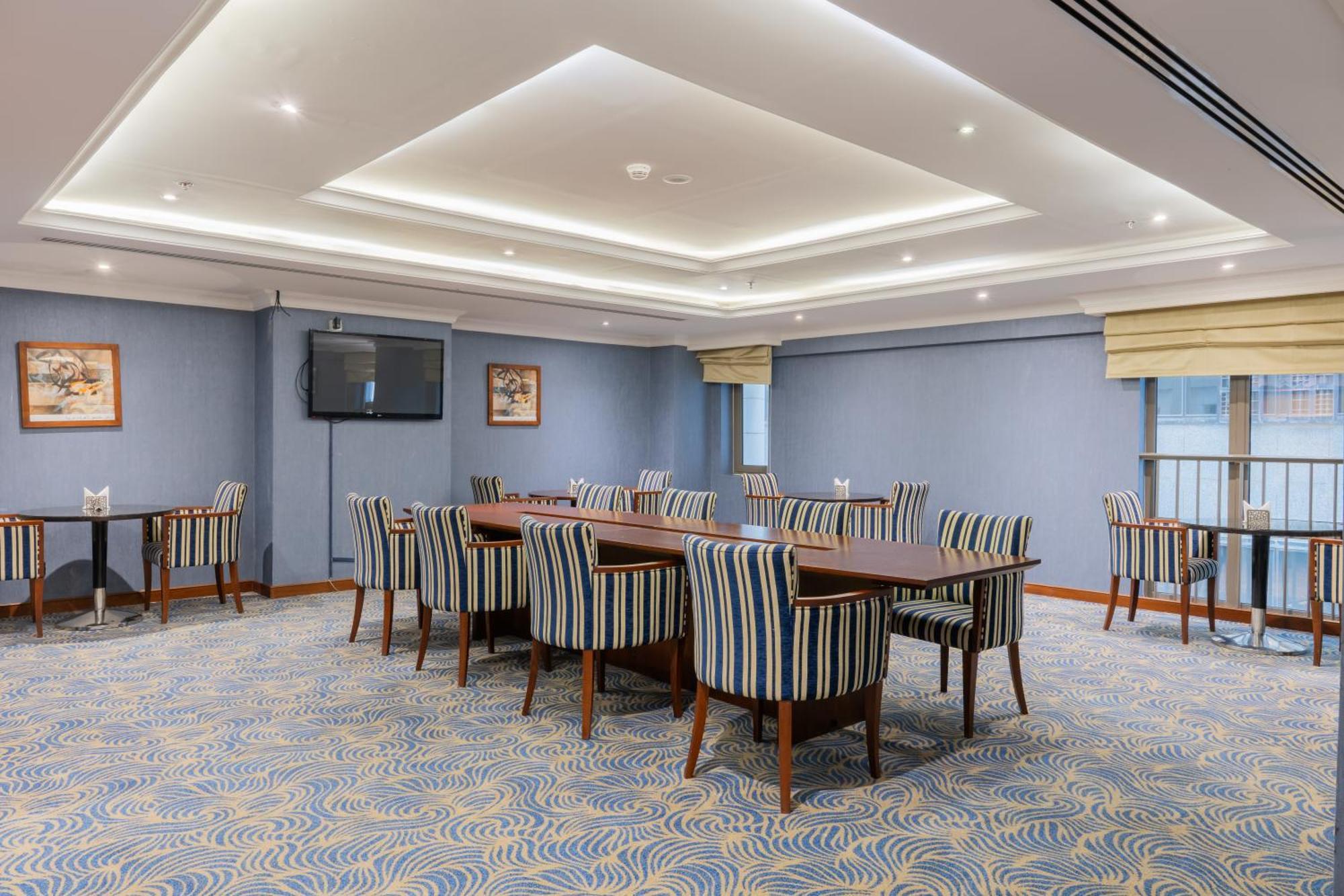 Al Aqeeq Madinah Hotel Медина Экстерьер фото The photo shows a spacious conference or meeting room. It features a light blue color scheme with a patterned carpet. There are several rectangular tables arranged in a group, surrounded by striped chairs. The room is well-lit with modern ceiling lig