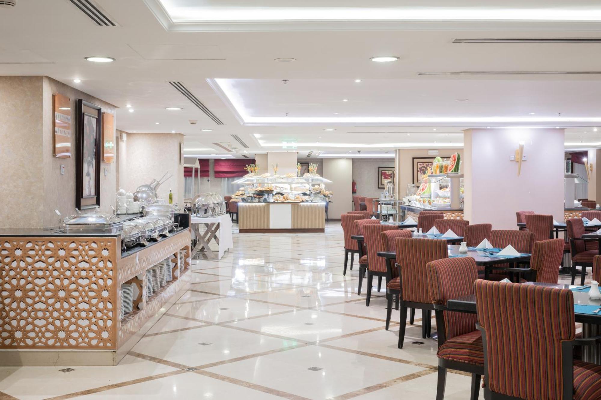 Al Aqeeq Madinah Hotel Медина Экстерьер фото The photo shows an elegantly designed restaurant or dining area. It features a spacious layout with a glossy tiled floor and bright, soft lighting. There are several dining tables with striped chairs arranged neatly. Along one side, there's a buffet 