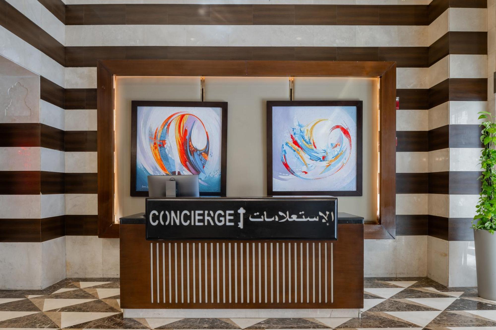 Al Aqeeq Madinah Hotel Медина Экстерьер фото The image shows a concierge desk in a hotel lobby. The desk is made of wood with a striped design and features two large, colorful abstract paintings on the wall behind it. The sign on the desk reads "CONCIERGE" in English and has Arabic text below i