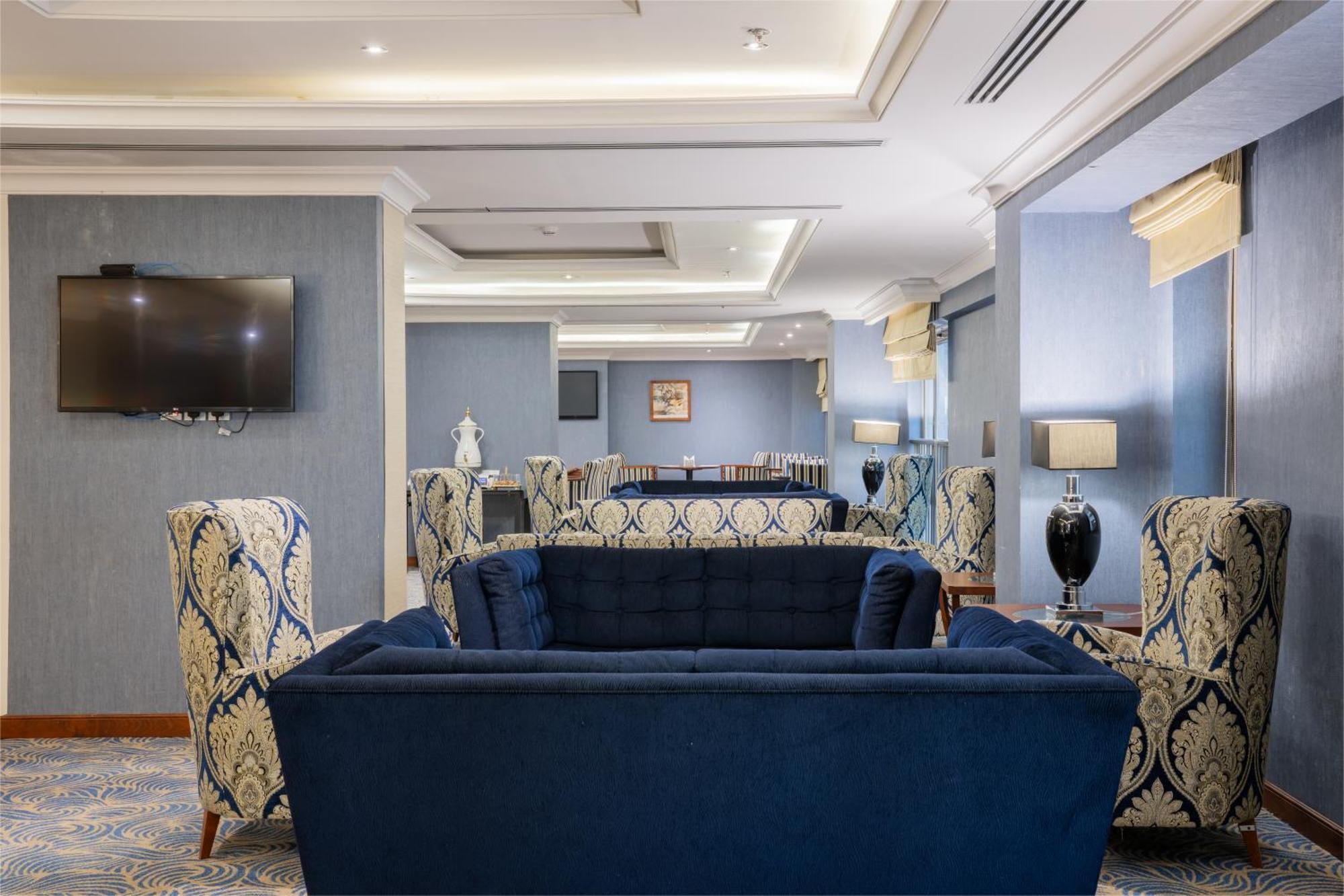 Al Aqeeq Madinah Hotel Медина Экстерьер фото The photo shows a stylish and modern lounge area. There are two patterned armchairs with blue and cream designs, positioned next to a deep blue couch. The room has a light blue wall with a flat-screen TV mounted on one side. Soft lighting from stylis