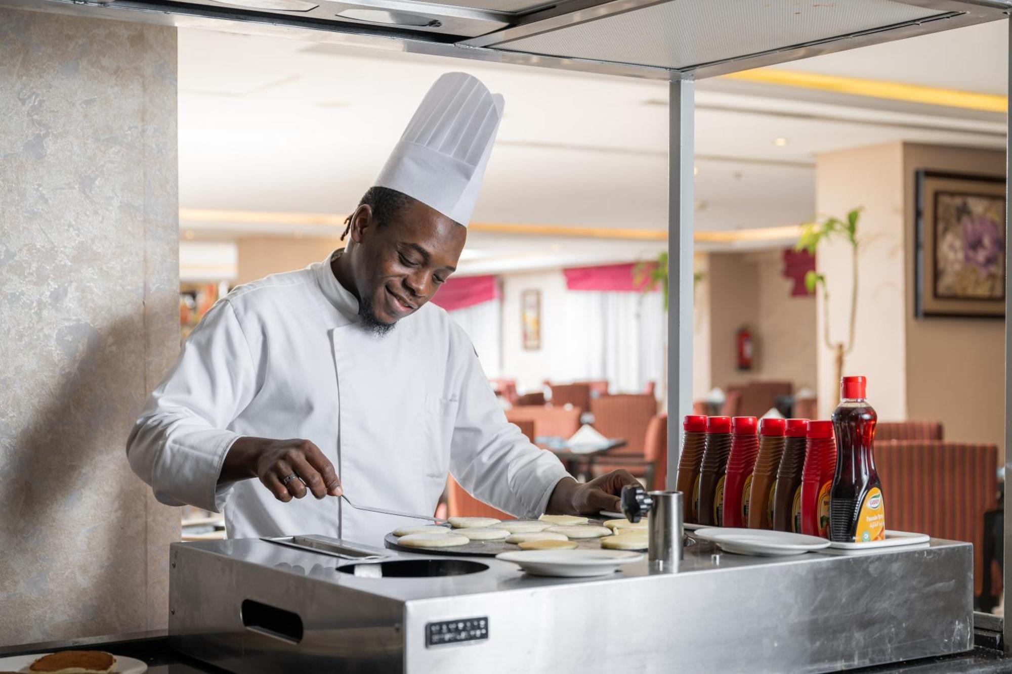 Al Aqeeq Madinah Hotel Медина Экстерьер фото The photo shows a chef in a professional kitchen setting. He is wearing a traditional white chef's uniform and a tall white chef's hat, and he appears to be smiling while cooking. The chef is standing at a stainless steel cooking station, where he is