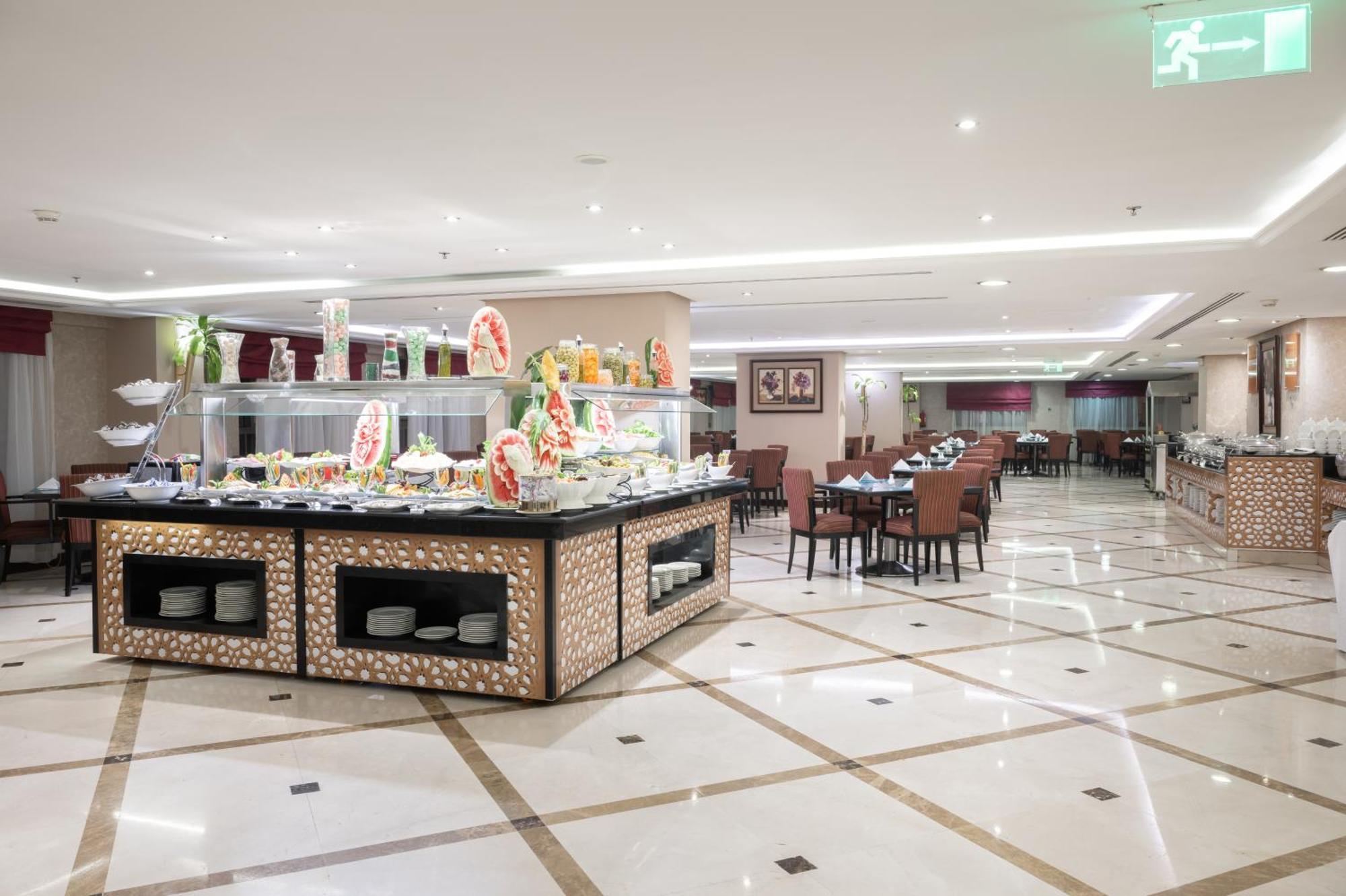 Al Aqeeq Madinah Hotel Медина Экстерьер фото The photo shows a spacious restaurant interior with a modern design. In the foreground, there is a large buffet counter filled with various food items, beautifully arranged and decorated. The counter features a backlit display, showcasing fruits and 