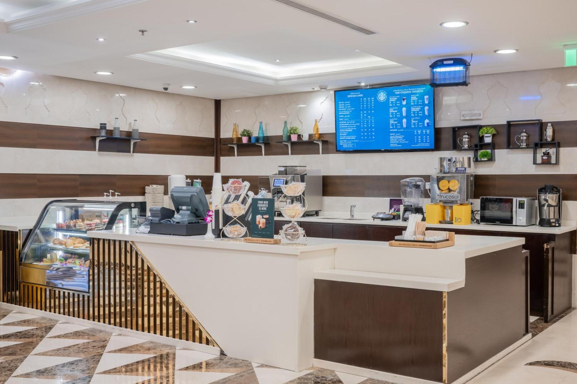 Al Aqeeq Madinah Hotel Медина Экстерьер фото The photo shows a modern café or coffee shop interior. There's a counter area made of smooth white and dark brown materials, featuring a cash register and display items. Behind the counter, shelves are stocked with various beverage items and equipmen