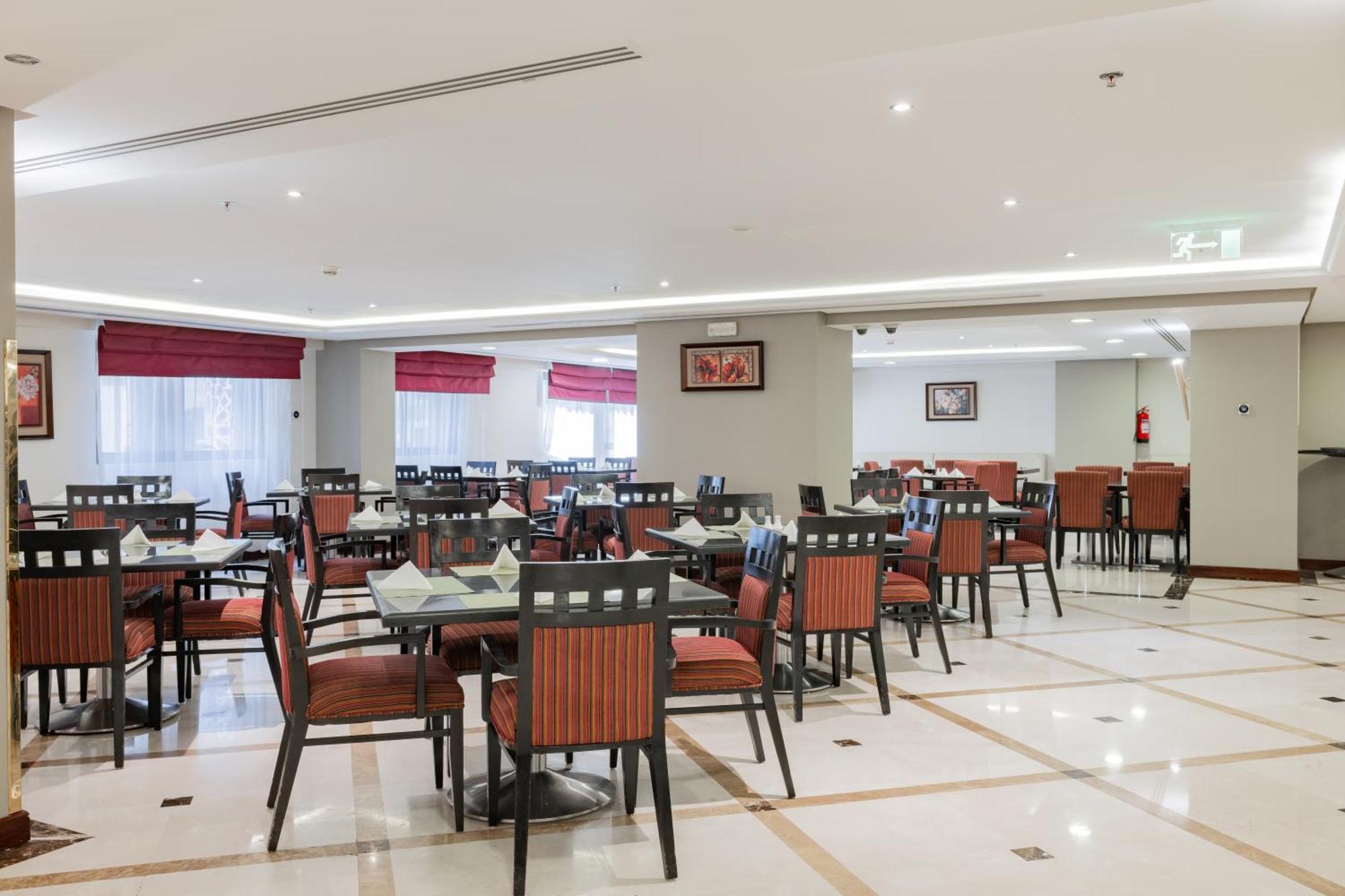 Al Aqeeq Madinah Hotel Медина Экстерьер фото The photo shows the interior of a restaurant or dining area. The space features several tables arranged with chairs, most of which have striped orange and black upholstery. The flooring is likely marble or tile, and the lighting is bright, creating a