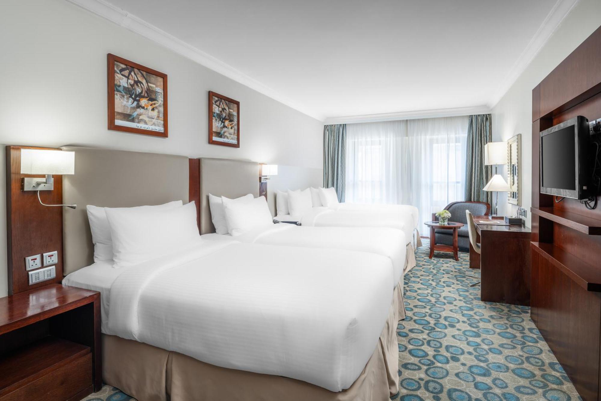 Al Aqeeq Madinah Hotel Медина Экстерьер фото The photo shows a hotel room featuring a modern, clean design. There are two large beds with white linens positioned side by side. Above the beds are framed artworks, and soft lighting is provided by wall sconces. To the right, there is a wooden ente