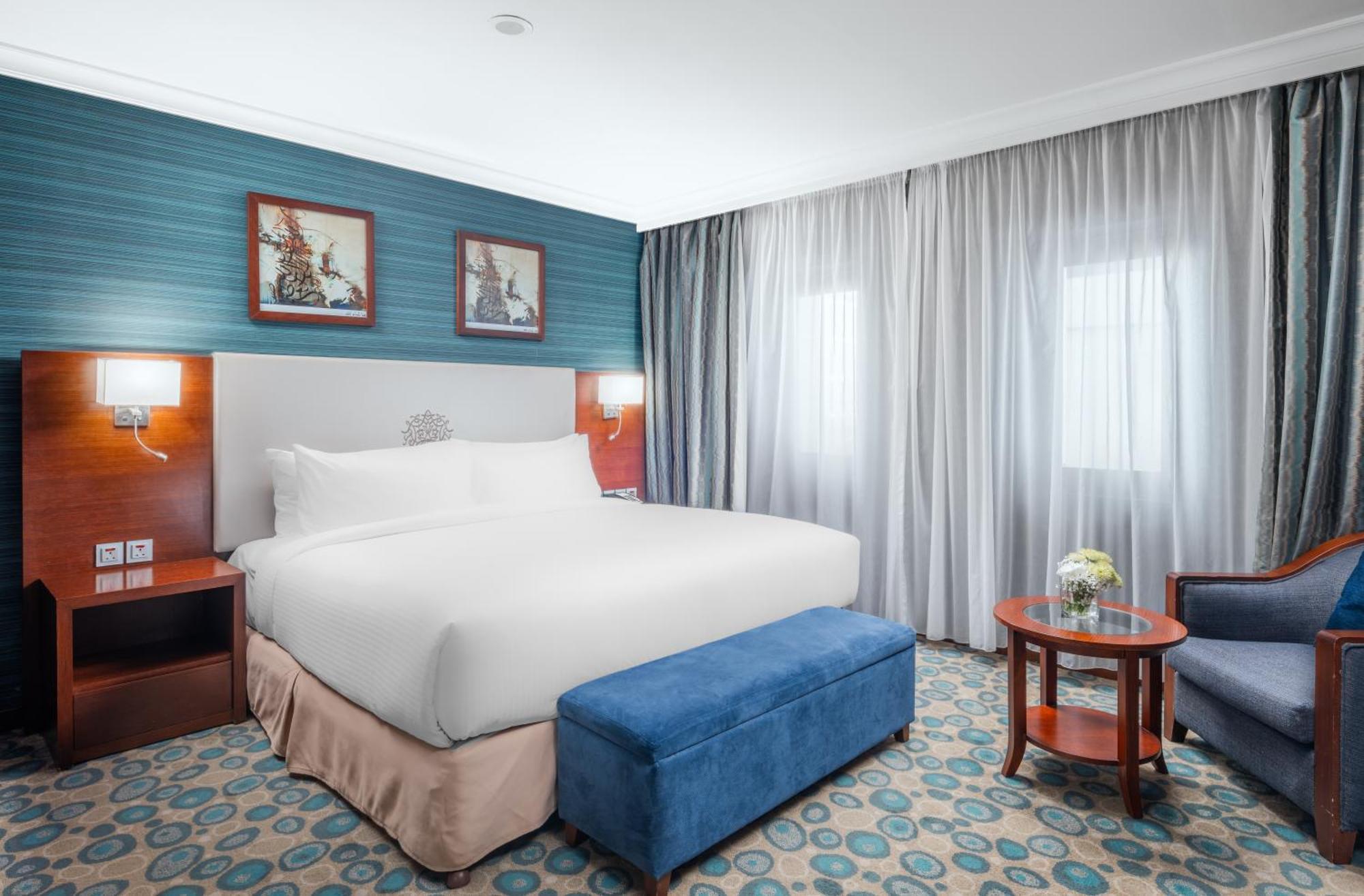 Al Aqeeq Madinah Hotel Медина Экстерьер фото The photo shows a hotel room featuring a large, neatly made bed with a white duvet. The walls have a light blue accent with framed artwork above the bed. There’s a blue ottoman at the foot of the bed. To one side, there’s a small wooden table with a 