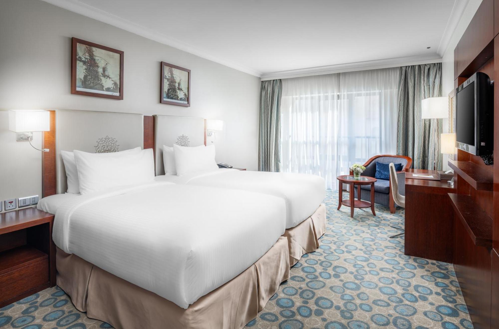 Al Aqeeq Madinah Hotel Медина Экстерьер фото The photo shows a hotel room featuring two white beds with plush bedding. There are decorative artworks on the walls above the beds. A large window with sheer curtains allows natural light into the space, illuminating the room. The floor is covered w