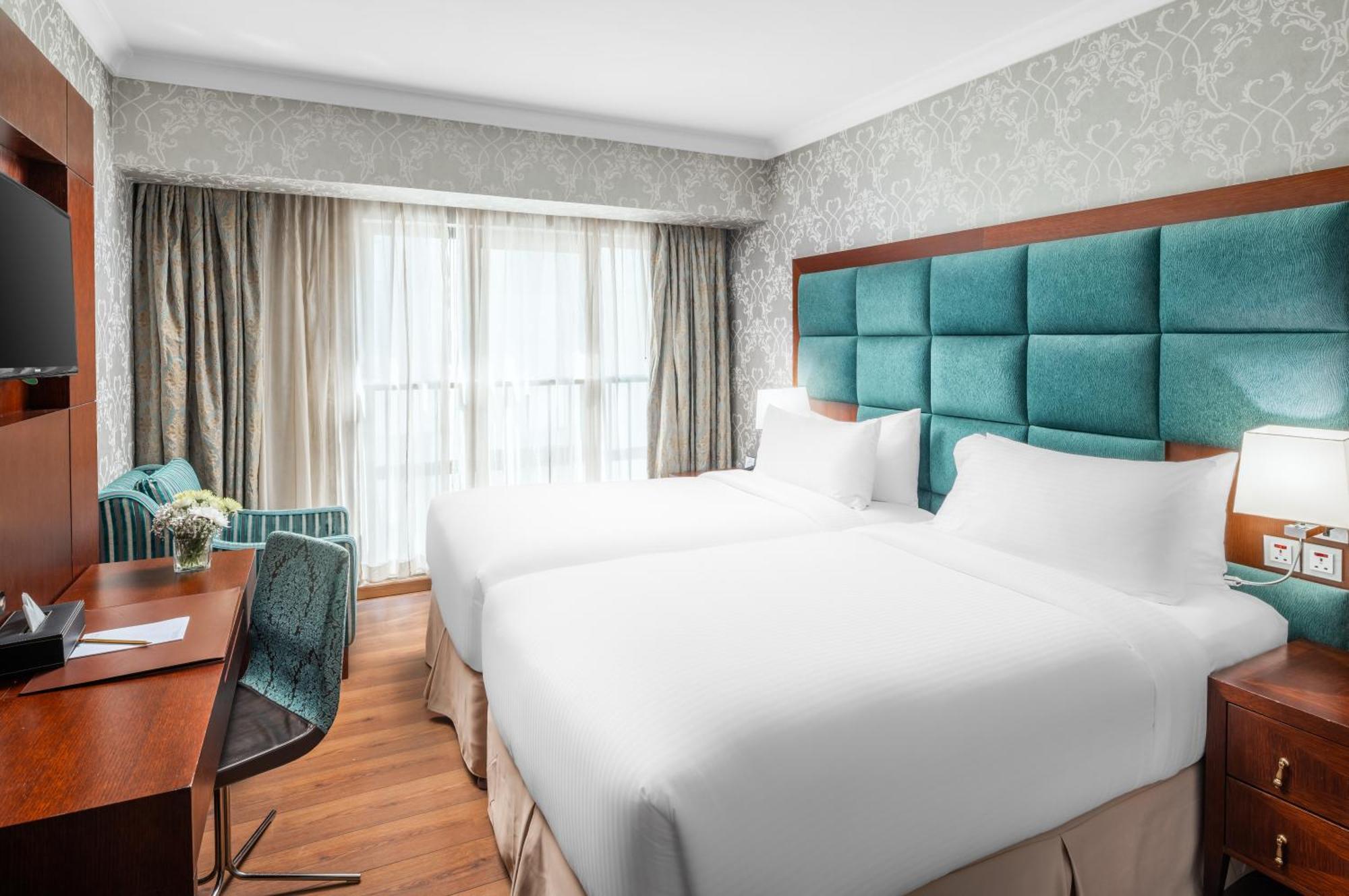 Al Aqeeq Madinah Hotel Медина Экстерьер фото The photo shows a modern hotel room featuring two single beds with white bedding. The headboard is upholstered in a teal fabric. There is a wooden desk and a chair near a window that is covered with sheer curtains and drapes. A cozy armchair in a mat