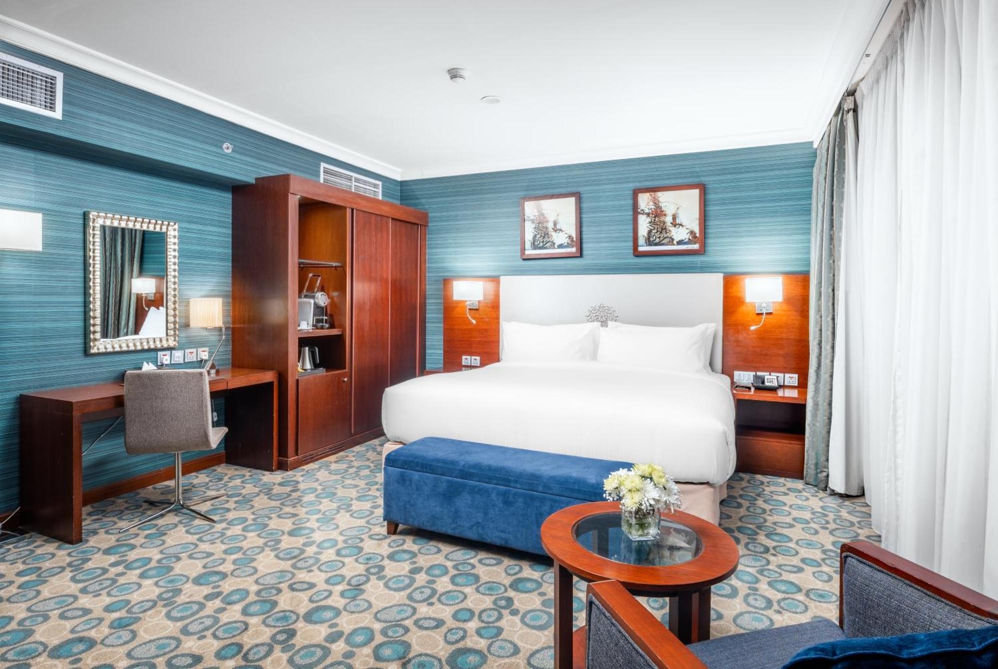 Al Aqeeq Madinah Hotel Медина Экстерьер фото The photo shows a well-decorated hotel room. It features a large bed with white bedding, flanked by wall-mounted lights. Behind the bed, there are two framed pictures hanging on a blue wall. To the left, there's a wooden wardrobe and a desk with a ch