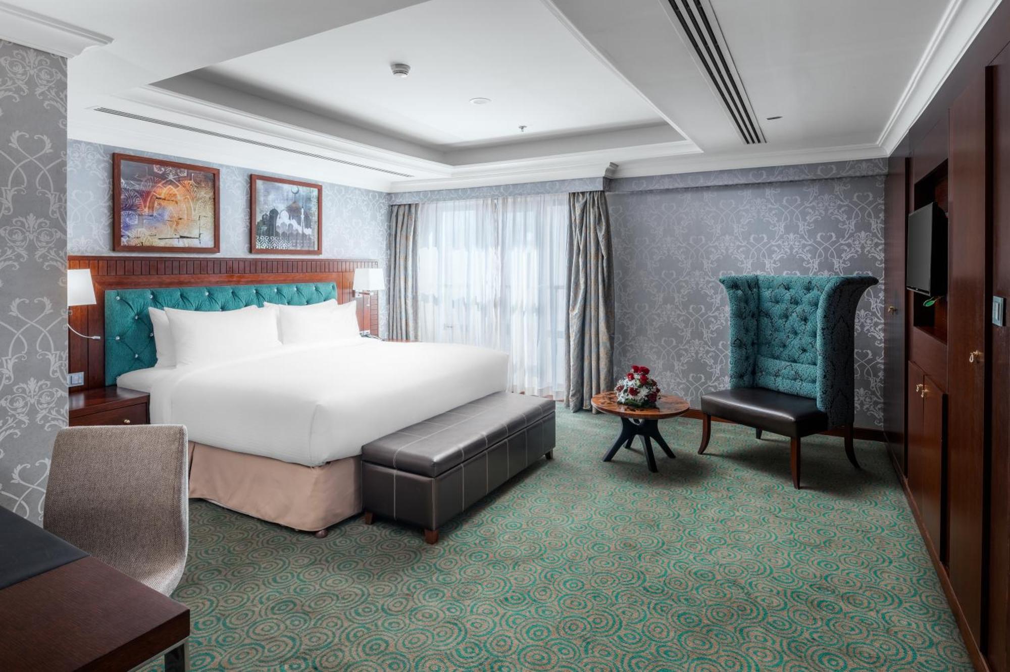 Al Aqeeq Madinah Hotel Медина Экстерьер фото The photo shows a well-appointed hotel room. It features a large bed with a tufted headboard in a turquoise color, complemented by decorative pillows. There is a comfortable ottoman at the foot of the bed and a small coffee table with a vase of flowe