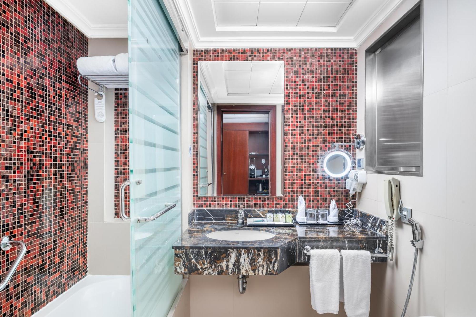 Al Aqeeq Madinah Hotel Медина Экстерьер фото The photo shows a modern bathroom featuring a mix of design elements. The walls have a vibrant red mosaic tile pattern, adding a bold touch to the space. There is a glass shower enclosure, and next to it, a sleek countertop with a dark, marbled surfa
