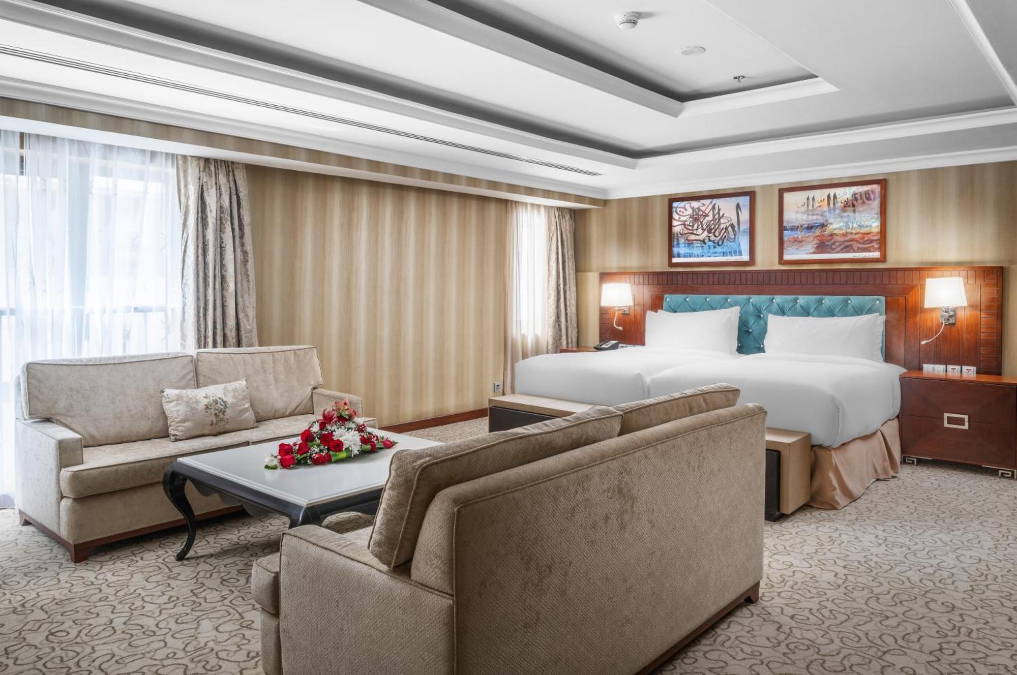 Al Aqeeq Madinah Hotel Медина Экстерьер фото The photo shows a well-decorated hotel room. It features a large bed with two pillows and a light-colored duvet. There are two comfortable couches arranged around a coffee table with a floral centerpiece. The walls have a subtle striped design, and l