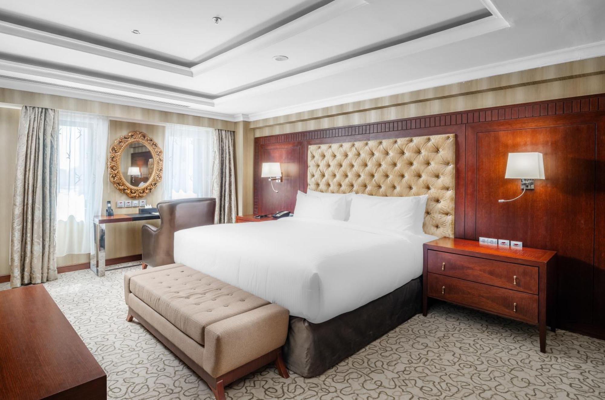 Al Aqeeq Madinah Hotel Медина Экстерьер фото The photo shows a well-furnished hotel room featuring a large bed with a tufted headboard. The room has a warm color palette with wooden accents and soft lighting. There are two windows with curtains that allow natural light to fill the space. Alongs