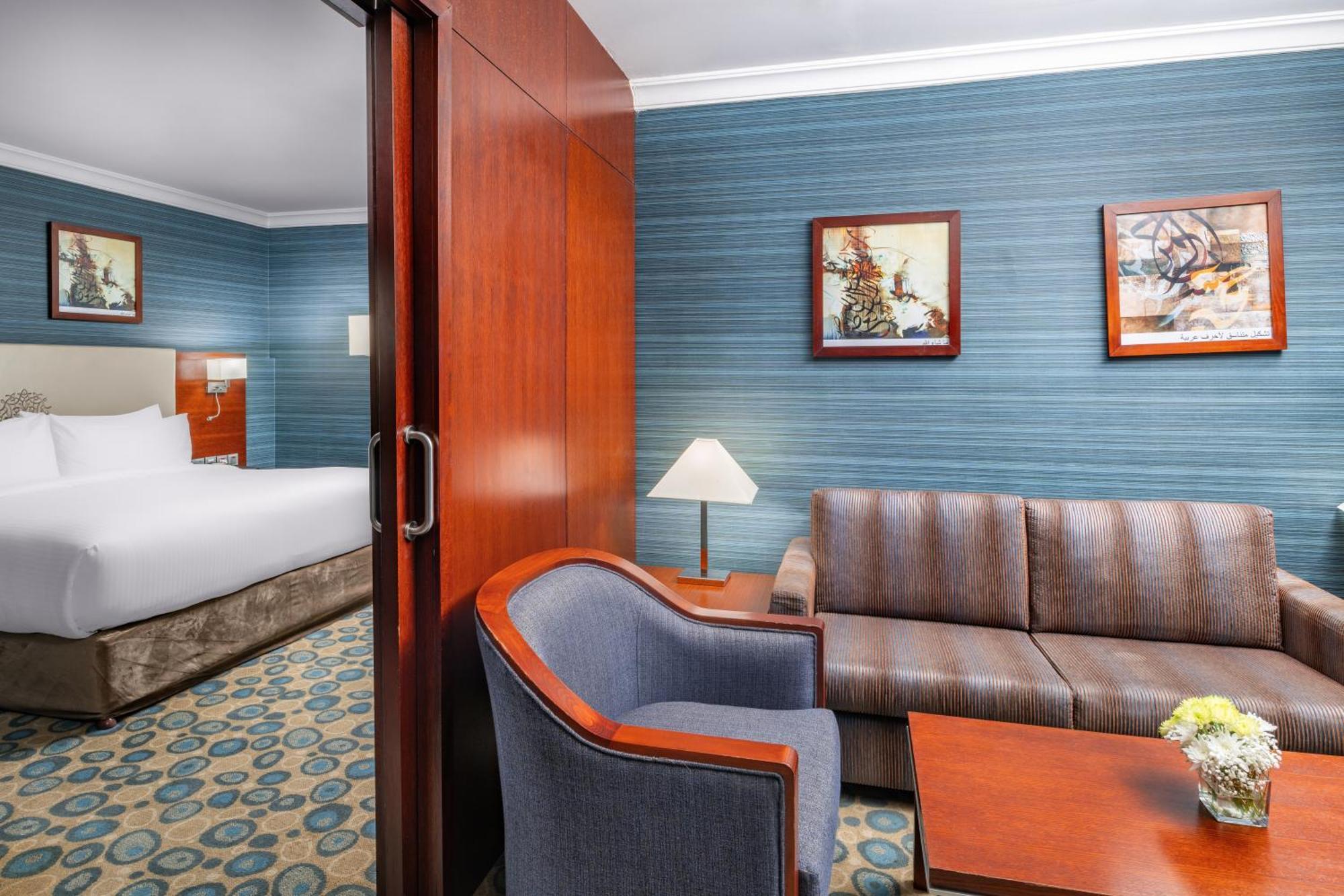 Al Aqeeq Madinah Hotel Медина Экстерьер фото The photo shows a well-furnished hotel room featuring a living area and a bedroom. To the left, there is a bed visible through an open doorway. The living area includes a gray upholstered chair and a brown couch, both arranged around a small table. A