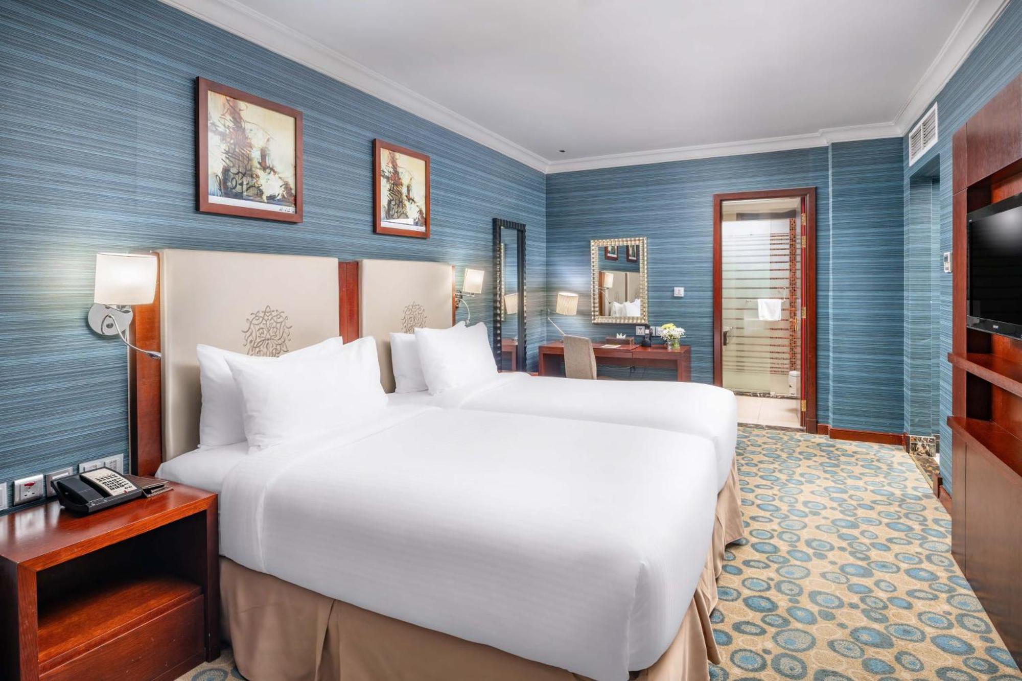 Al Aqeeq Madinah Hotel Медина Экстерьер фото The photo shows a hotel room featuring two twin beds with white bedding, positioned against a light-colored headboard. The walls are painted in a soft blue shade, adorned with framed artwork. There is a wooden desk with a chair, and a mirror mounted 