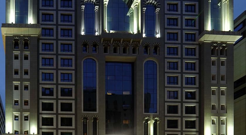 Al Aqeeq Madinah Hotel Медина Экстерьер фото The image shows the facade of a modern high-rise building illuminated at night. The design features large glass windows arranged in a grid pattern, along with decorative architectural elements at the top, including arches and ornate detailing. The co
