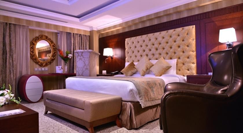 Al Aqeeq Madinah Hotel Медина Экстерьер фото The photo shows a luxurious hotel room. It features a large, upholstered headboard with a tufted design, paired with plush bedding and decorative cushions. There's a stylish bench at the foot of the bed and a comfortable armchair to the side. The roo