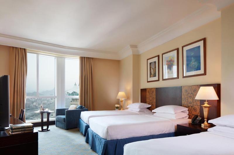 Al Aqeeq Madinah Hotel Медина Экстерьер фото The photo shows a hotel room featuring three beds arranged neatly. The room has a warm, inviting ambiance with beige walls and a soft carpet. Large windows allow natural light to fill the space, offering a view outside. There are curtains in a light 