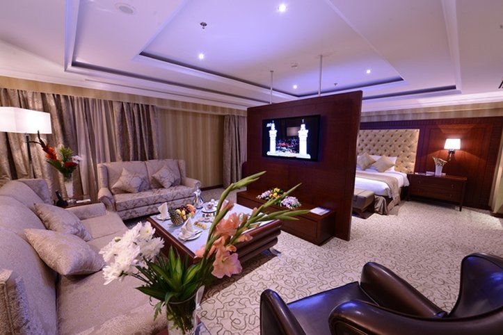 Al Aqeeq Madinah Hotel Медина Экстерьер фото The photo shows a luxurious hotel room suite. There is a comfortable seating area with stylish sofas and a coffee table. A decorative arrangement of flowers is on the table, adding a touch of elegance. A large flat-screen television is mounted on a p