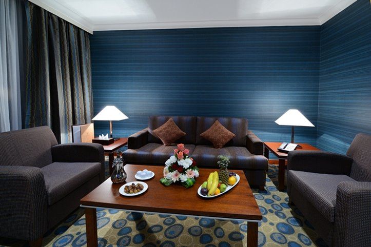 Al Aqeeq Madinah Hotel Медина Экстерьер фото The photo shows a cozy living area featuring a dark blue wall with a textured design. In the center, there is a wooden coffee table surrounded by two comfortable armchairs and a sofa with decorative cushions. On the table, there are plates with snack