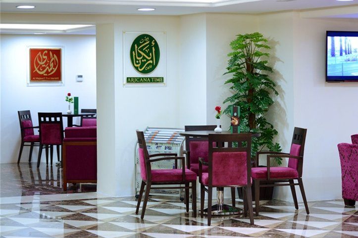 Al Aqeeq Madinah Hotel Медина Экстерьер фото The photo shows an interior space, likely a dining or lounge area. There are several tables with four chairs each, featuring a modern design with dark wood and plush maroon upholstery. On the walls, there are decorative signs, one of which has Arabic