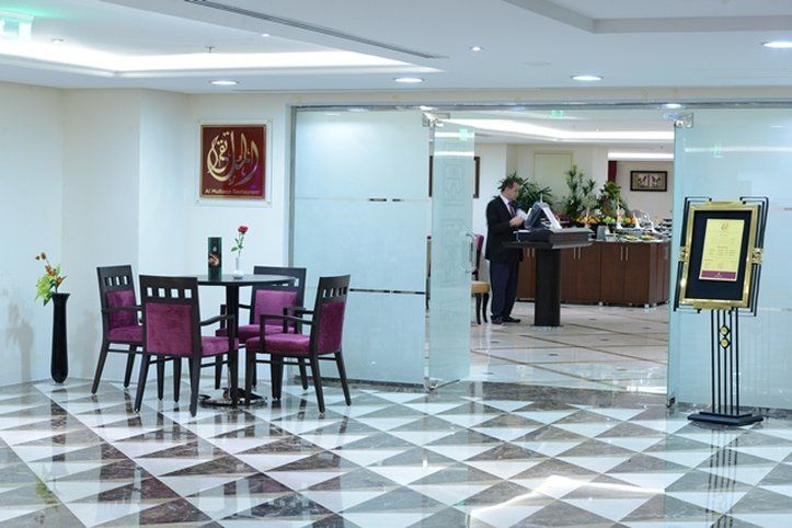 Al Aqeeq Madinah Hotel Медина Экстерьер фото The photo shows a modern interior space, likely a lobby or reception area of a hotel or restaurant. It features a checkered floor with a combination of dark and light tiles. On the left, there is a small table set for two, adorned with a single rose 