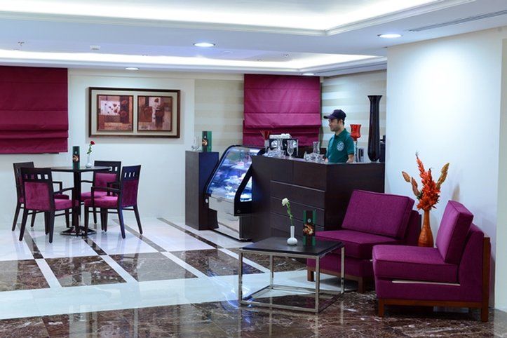 Al Aqeeq Madinah Hotel Медина Экстерьер фото The photo shows a modern interior space, likely a café or small restaurant. It features a seating area with a few tables and chairs, all in a dark wood finish with purple upholstery. There is a counter area with a staff member behind it, possibly ser