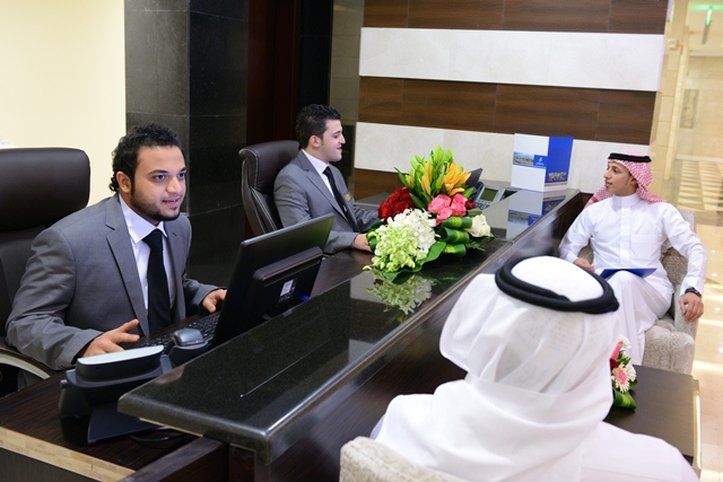 Al Aqeeq Madinah Hotel Медина Экстерьер фото The photo depicts a reception area with three individuals. Two men are seated behind a reception desk; they are dressed in formal suits and appear to be engaged in conversation, possibly assisting a guest. The desk has a computer and a vibrant arrang